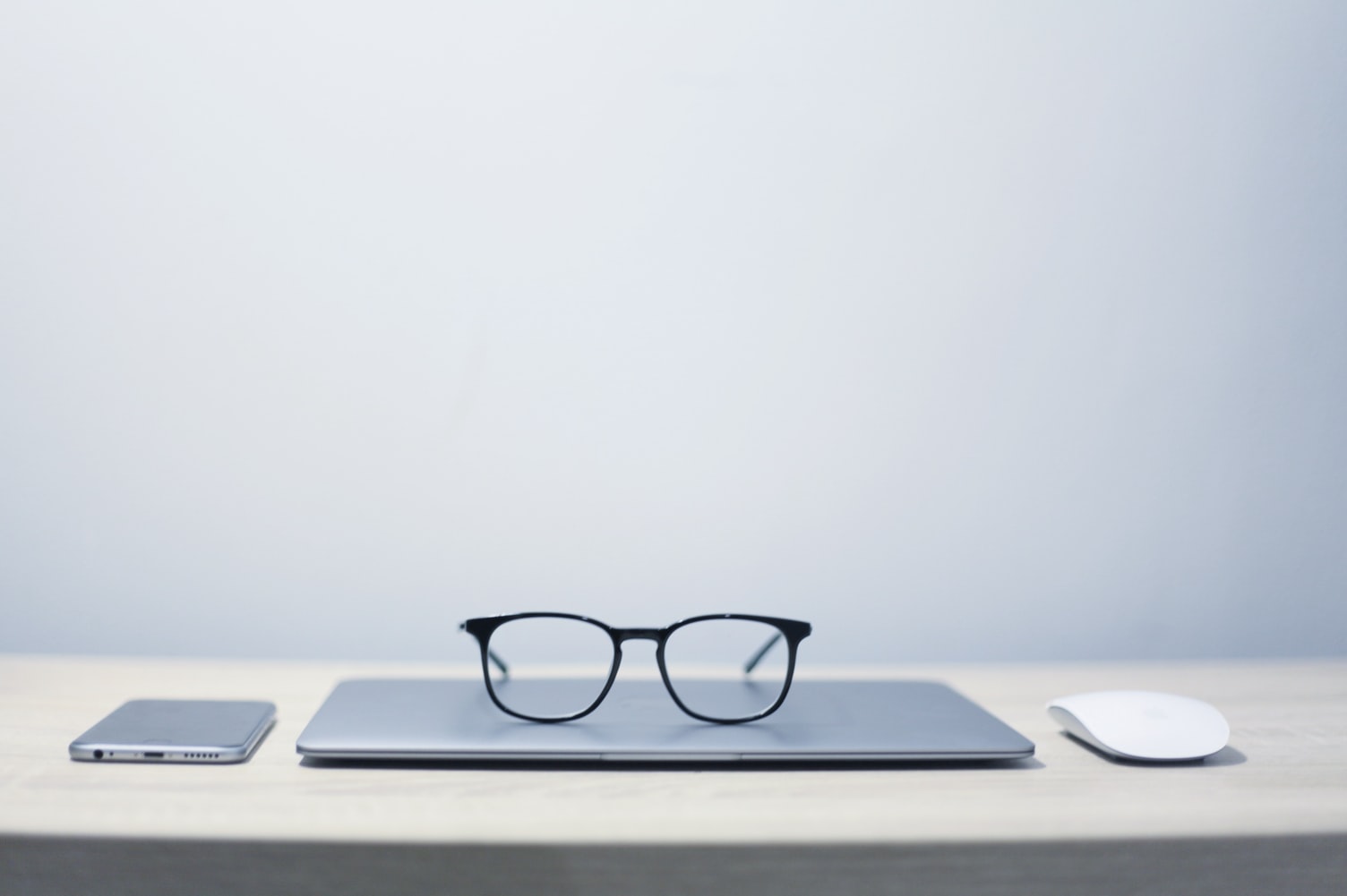 Photo of a desktop containing a mobile phone, eyeglasses, a closed laptop computer, and a mouse.