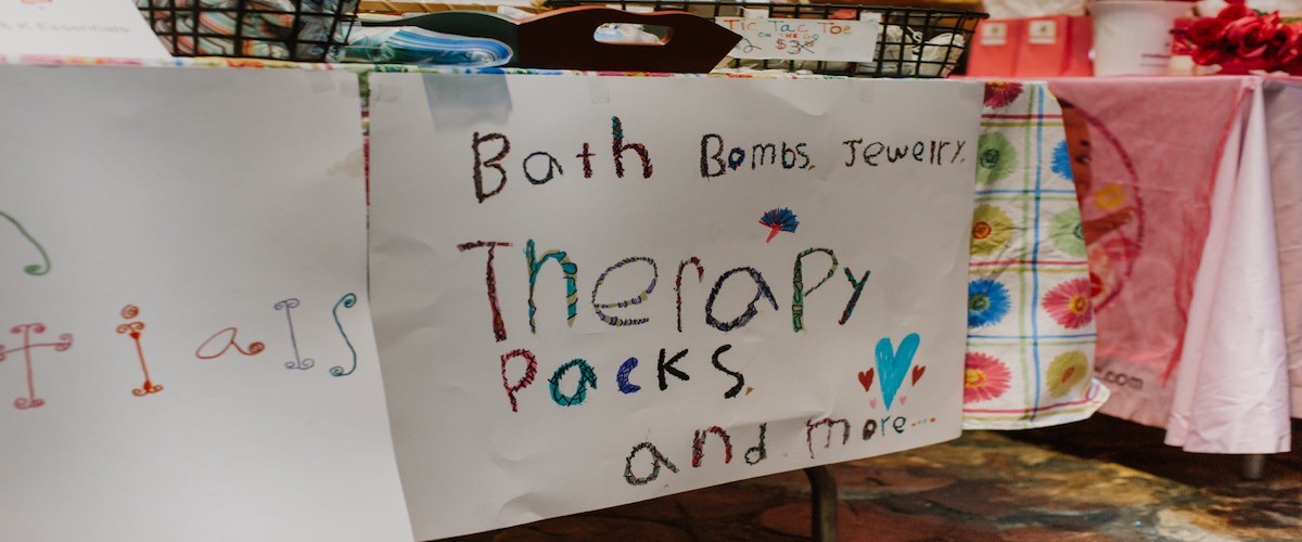 Photo of a sign written in crayon by a child’s hand that says, “Bath bombs, Jewelry. Therapy packs and more”.