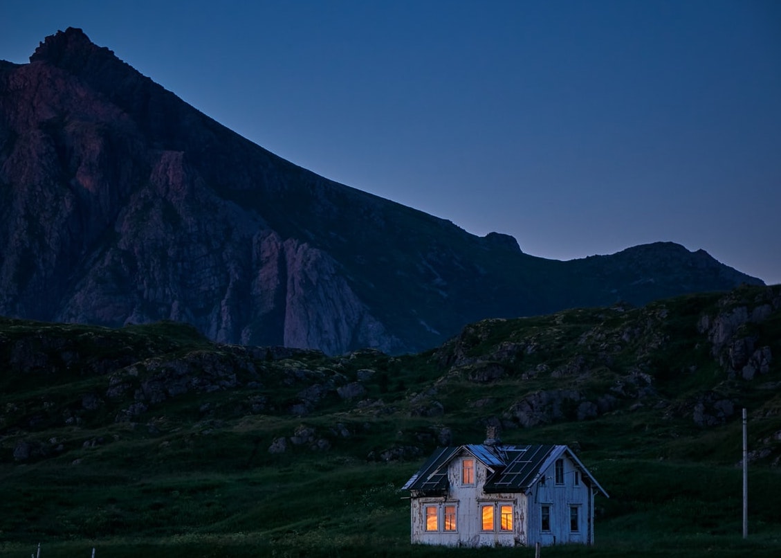 Photo of a small house in the mountains at dusk. Lights can be seen illuminated via all the windows in the front of the home.